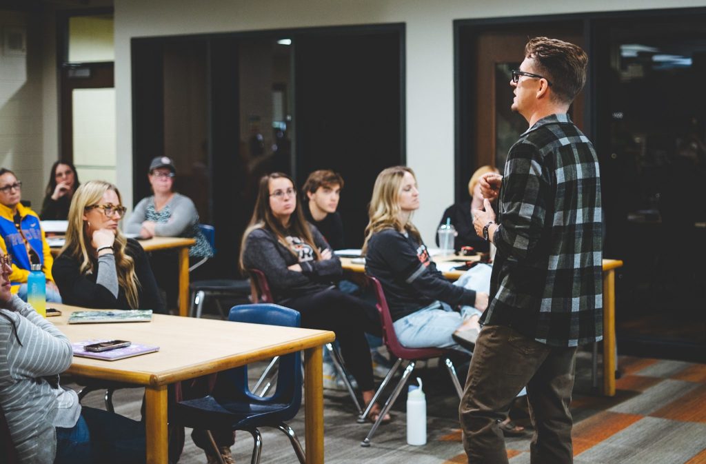 A speaker engages an audience in a classroom setting, with attendees seated at tables listening attentively.